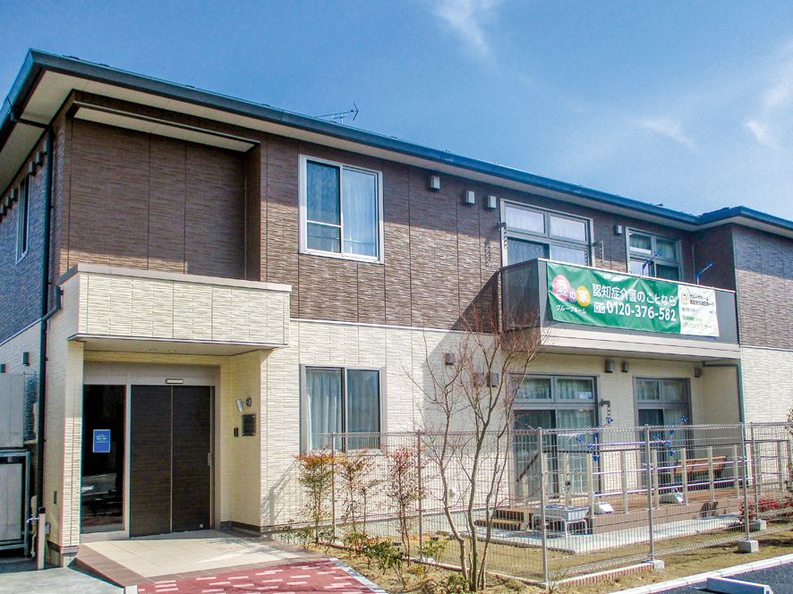 ２階建てのブラウン系の建物には玄関に屋根とスロープがあり、木もたくさん植えられている。