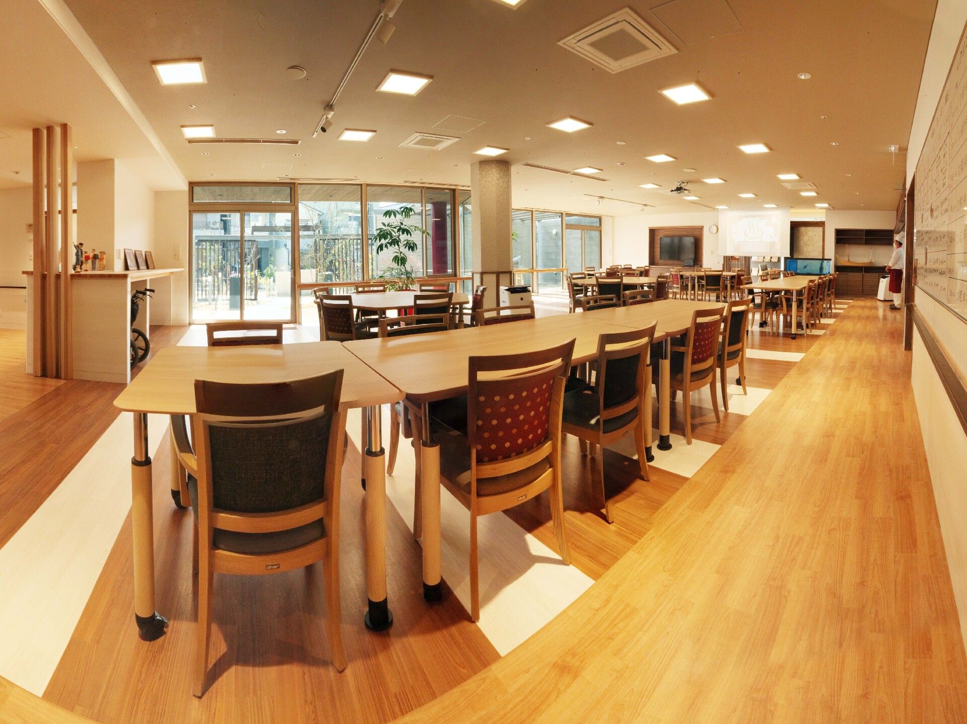庭園を見渡すことができる大きな窓が付いた食堂には大小のテーブルセットが複数置かれ、左奥の棚には車いすも収納されている。