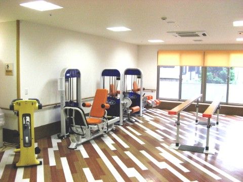 部屋の中央に歩行訓練機器があり、壁面沿いにトレーニングマシーンが数台並んで設置されているトレーニングルーム