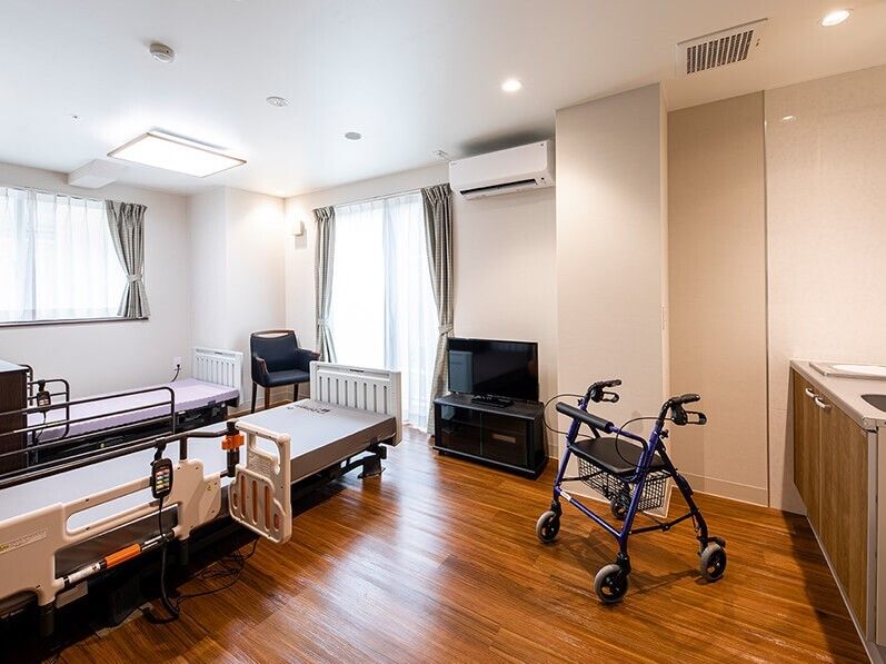 2台のベッドが並べられたフローリングの部屋にはキッチンもあり、壁にはエアコンが付き、テレビやいすも置かれている。