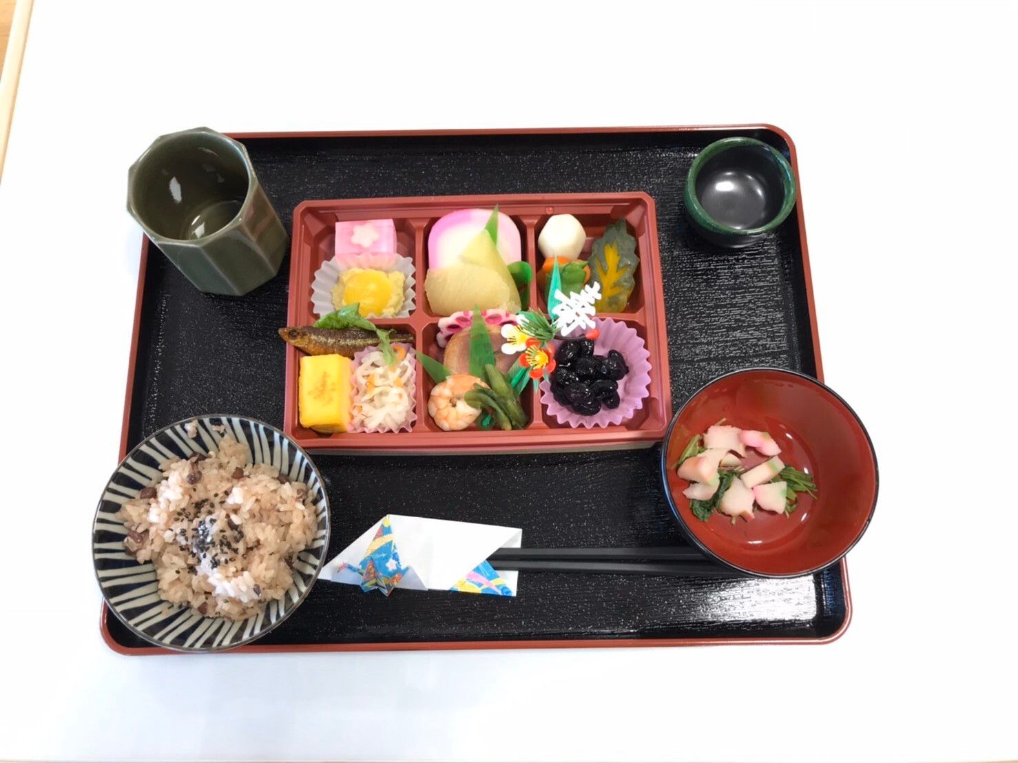６つに仕切られた弁当風の容器に黒豆や紅白のかまぼこ、エビなど正月料理が並び、赤飯と吸い物が添えられている。