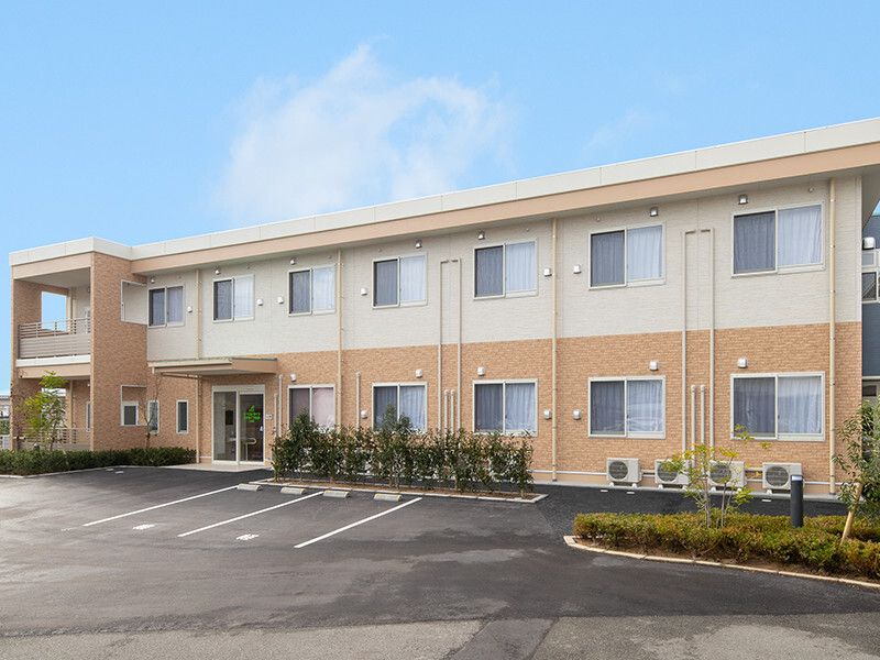 ベージュと白の２階建ての建物は横に長く、敷地内には駐車場がある。草木も植えられている。
