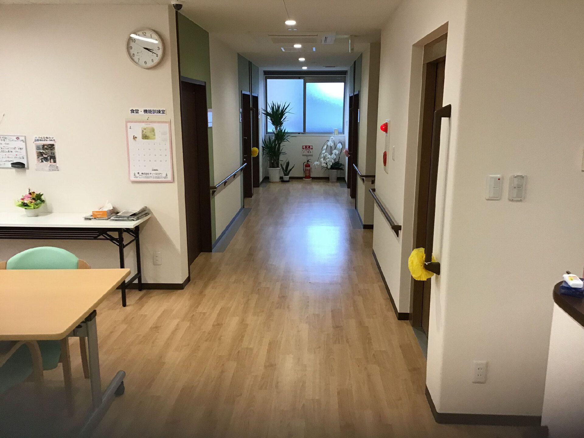 まっすぐな廊下の突き当りの窓の下には花や草木が飾られ、手前にはダイニングテーブルセットが置かれている。