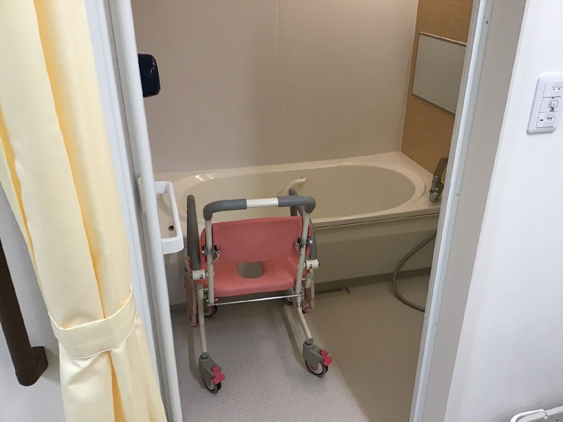 ピンクの車いすタイプの椅子が設置された浴室には一人用の浴槽が設置され、シャワーも付いている。