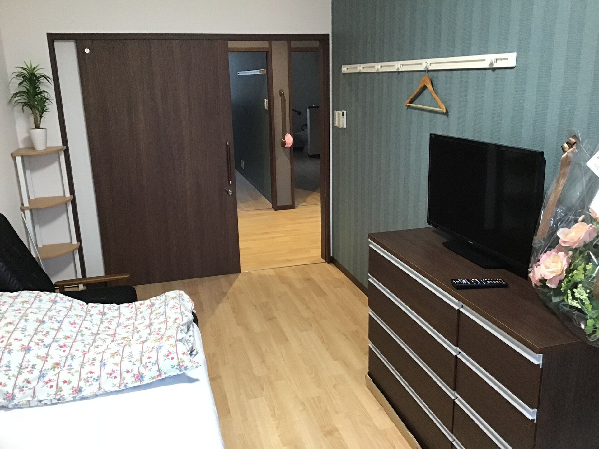 寝具付きのベッドやタンスがあり、タンスの上には花やテレビが置かれている。フローリングの部屋。