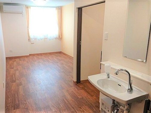車いす対応の洗面所の隣にはトイレのスライド式のドアが付き、エアコンやカーテンが付いたフローリングの部屋。
