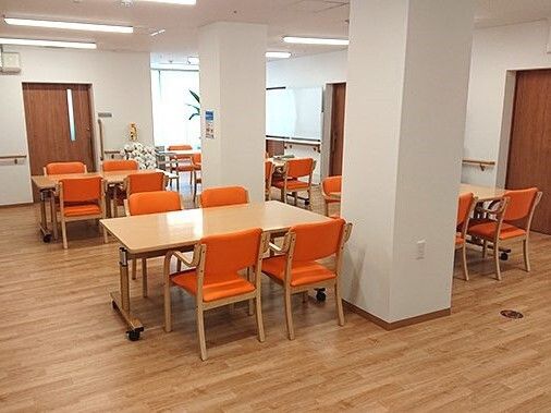 オレンジの椅子がセットされたダイニングテーブルが複数置かれた明るい食堂。観葉植物や花も飾られ、親しみやすい雰囲気。