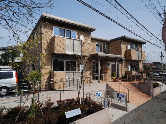 2階建ての建物。ブラウン系で玄関に屋根が付き、建物周辺には手すり付きの階段とスロープがある。