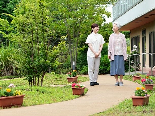 パジャマ姿でベッドに横になっている高齢女性に対し、笑顔で介護に当たるエプロン姿の女性スタッフ。