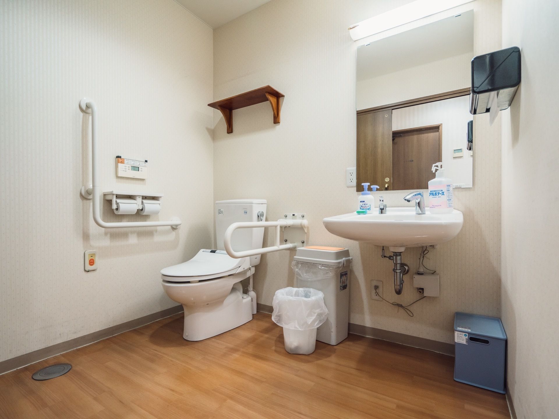 トイレの床はフローリングで、便座の横と壁には手すりが設置されている。便座の右側にはゴミ箱や洗面台がある。