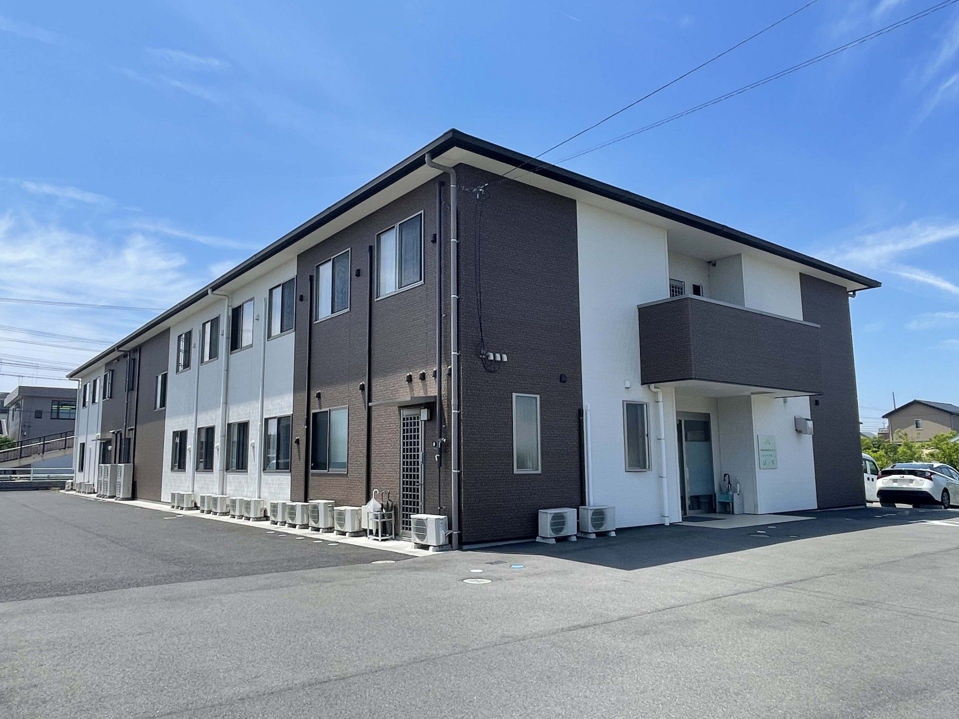 2階建ての建物はブラウンと白で落ち着いた雰囲気。建物周辺には緑も植えられている。