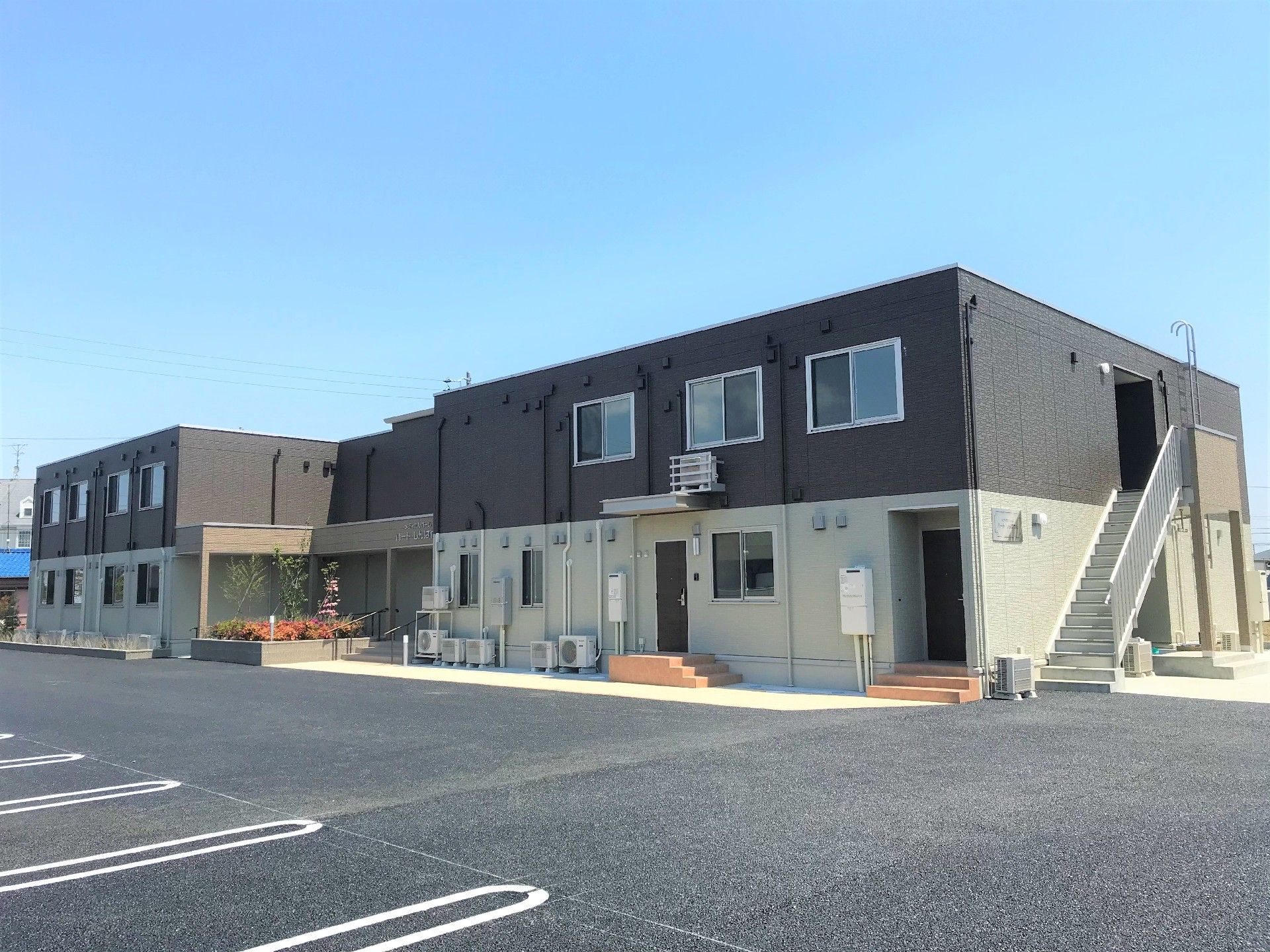 広い駐車場がある敷地にモノトーン系の建物が建っている。建物の側面には屋外階段もある。