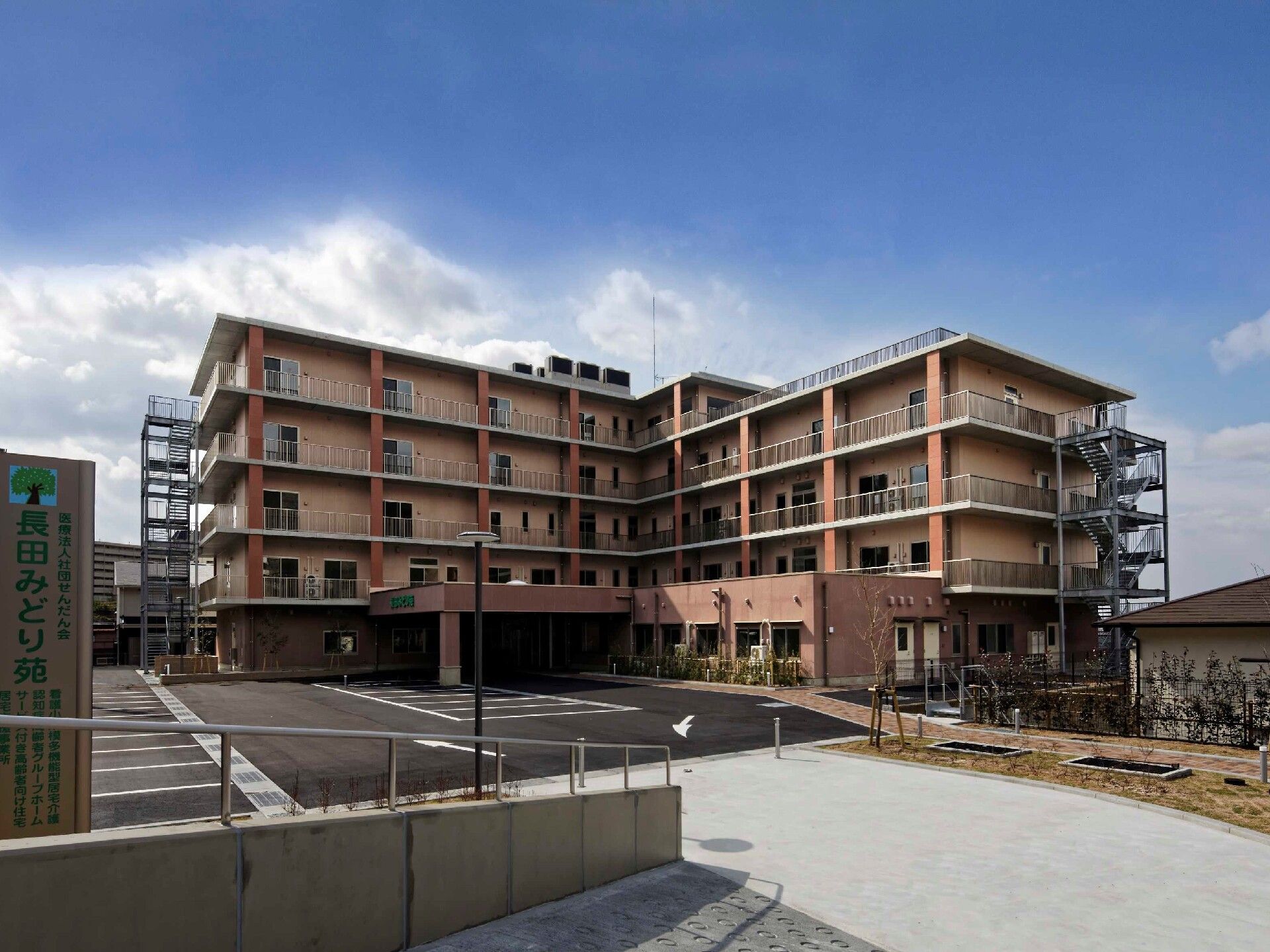 ベージュ系の建物はエル字型で大きな駐車場が付き、半分4階建て、もう半分が5階建てとなっていて、周囲には青い空が広がっている。