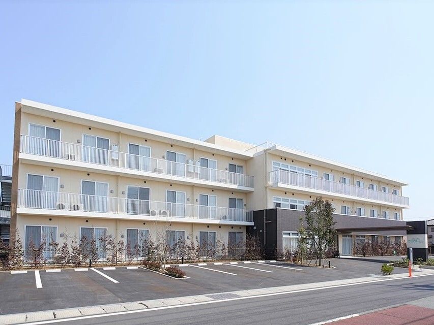 駐車場が付いた白っぽい３階建ての建物には玄関に大きな車寄せが付き、周辺には緑もある。