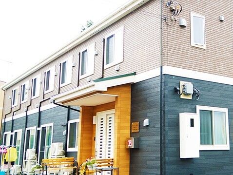 ブラウンとグリーンの2階建ての建物。建物の前には洗濯物干し場があり、玄関前にはベンチが並んでいる。