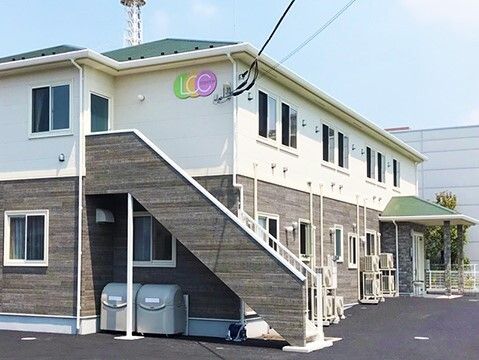 1階部分は木目調、2階と3階は白で、周辺には緑があり、敷地内にはゴミ置き場や駐車スペースもある。