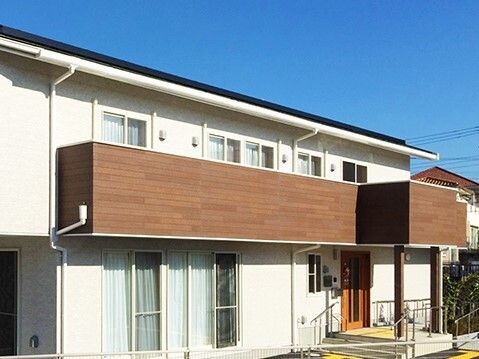 ベランダのついた2階建ての建物は白とブラウンで、玄関部分に手すり付きの階段とスロープが付いている。