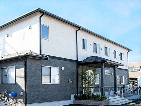 玄関には手すり付きのスロープと階段があり、緑も植えられている。2階建ての建物はグレーと白のモノトーン。