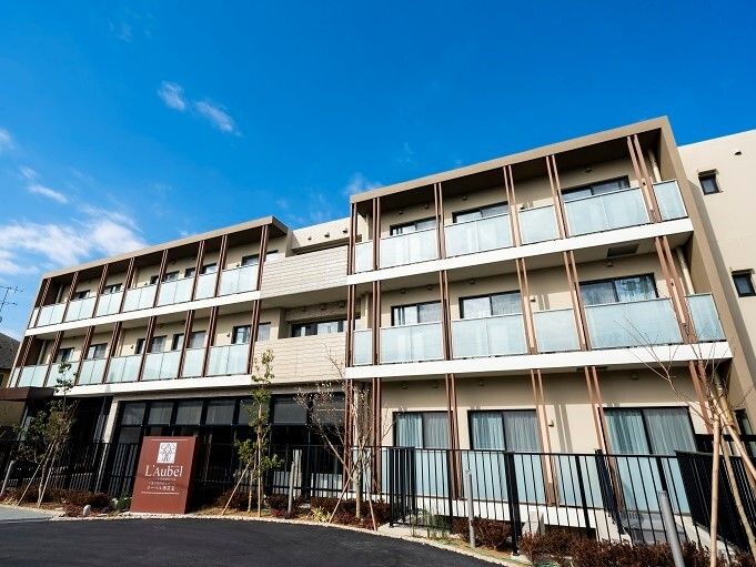 各フロアにベランダが付いた3階建ての建物。建物の前にはブラウンの館銘板があり、建物の周辺には黒いフェンスも建てられている。