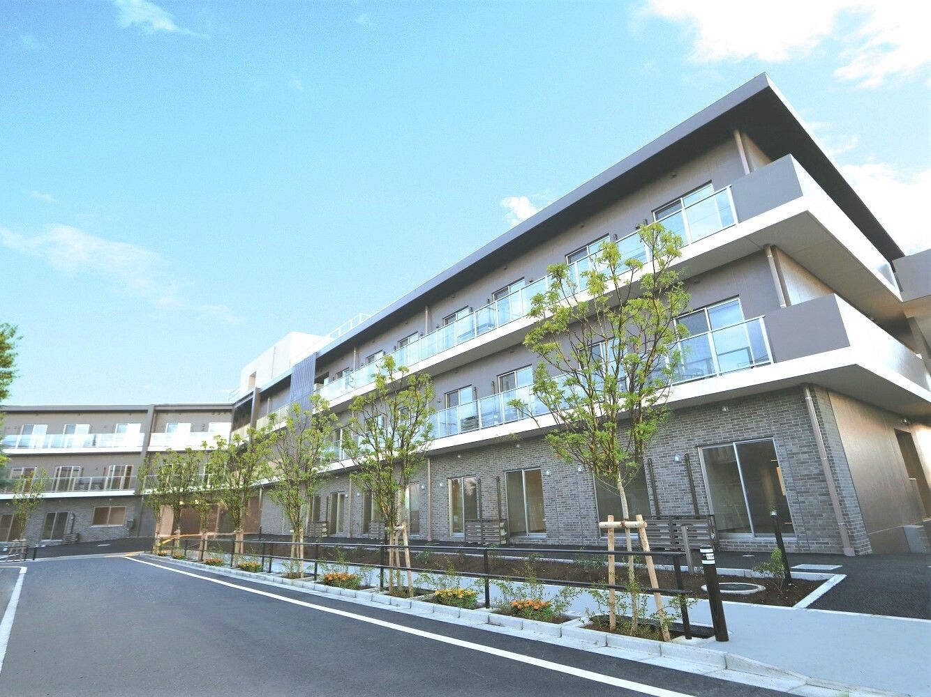 木や花が植えられた道路沿いに建つ、三階建てのベランダが並んだ建物