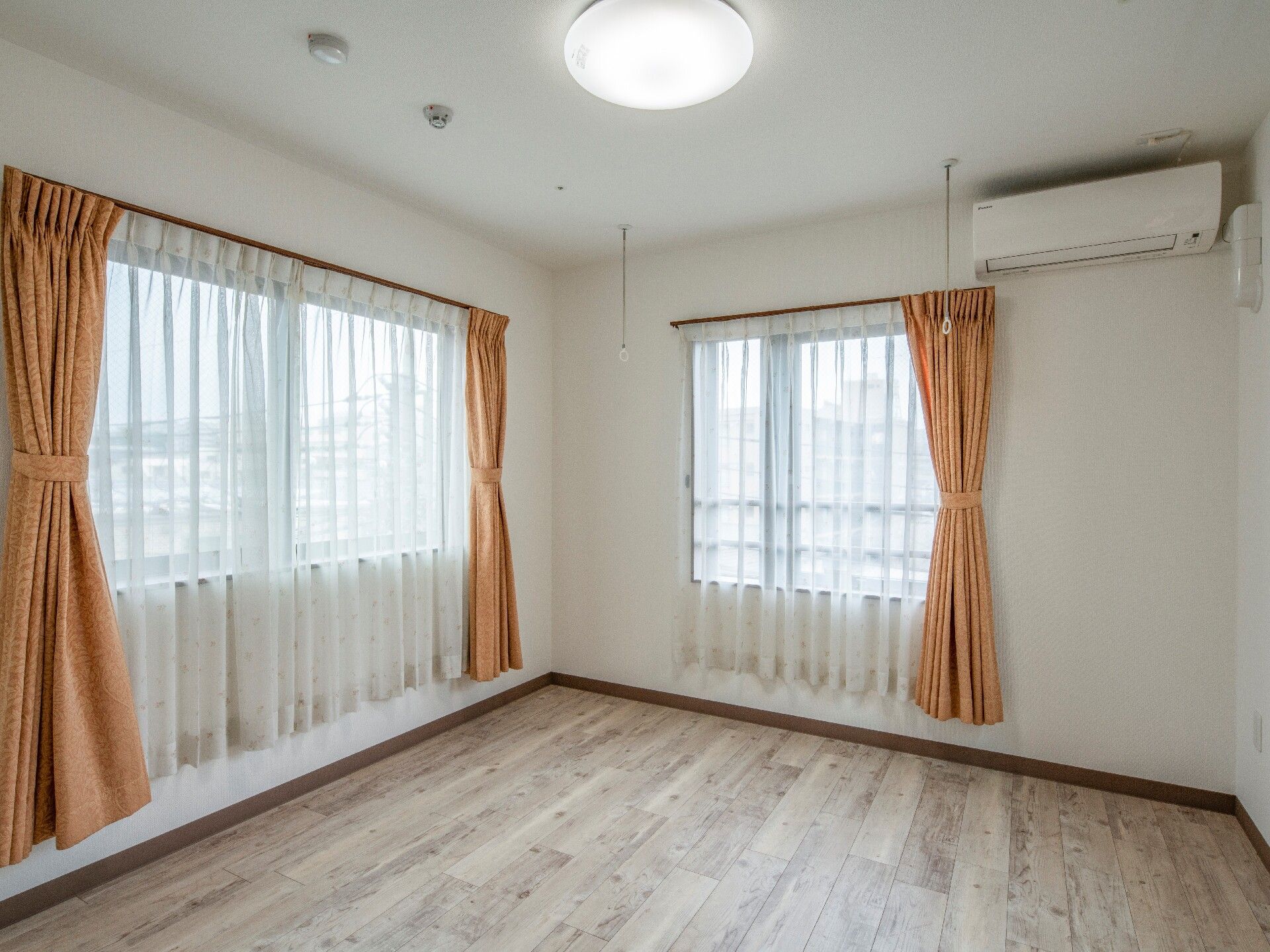 レースカーテンとカーテンが付いた大きな窓が２つある広いフローリングの部屋。明るい雰囲気で、壁にはエアコンがあり、天井の照明は電灯が点いている。
