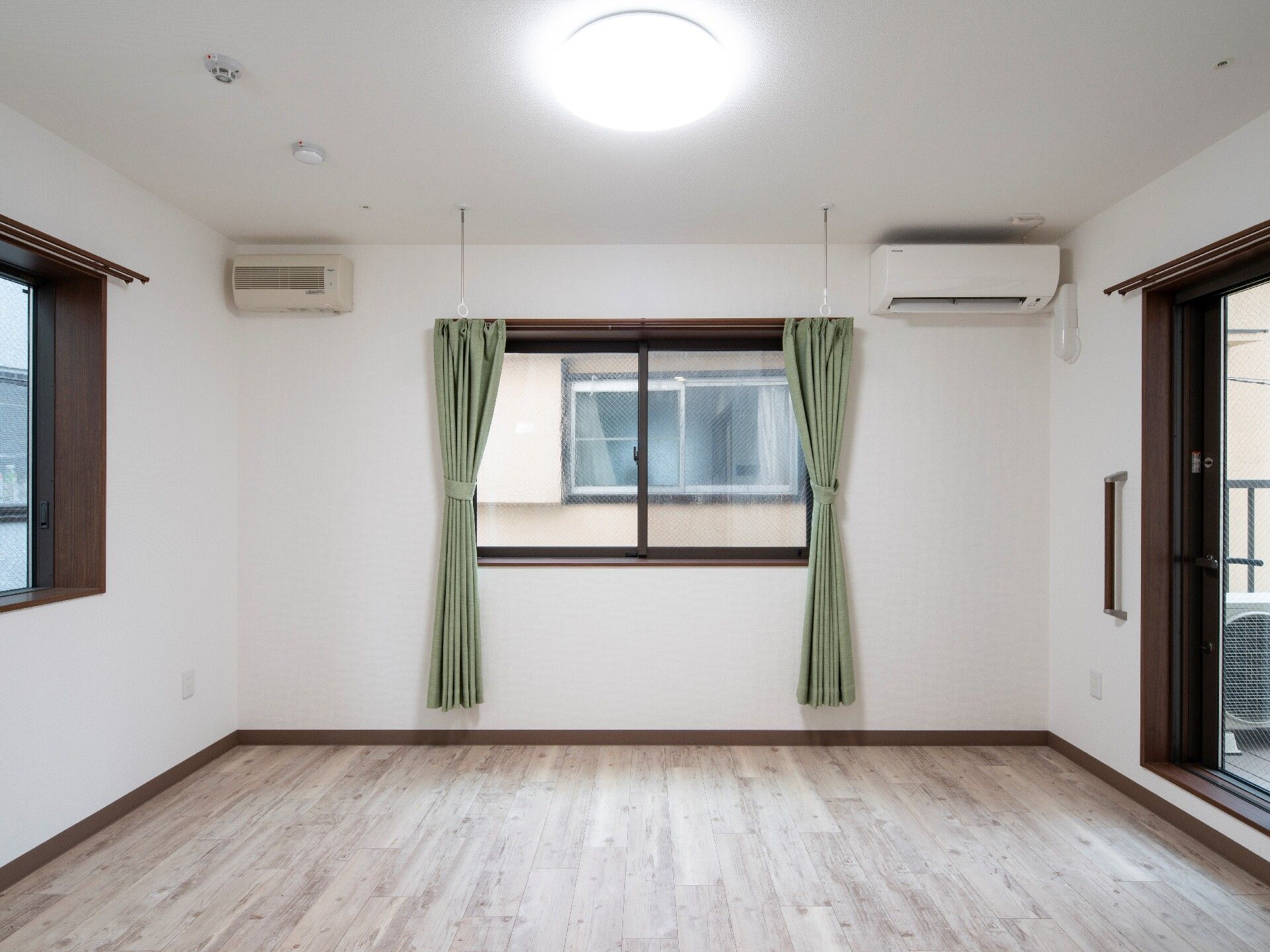 正面にある窓には竹が長めの緑のカーテンが設置され、窓の外にはほかの建物の窓が見える。右側のベランダにつながっている窓のそばには手すりがある。