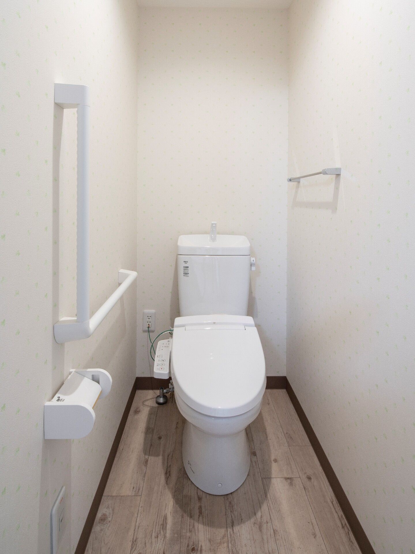 白いエル字型の手すりが設置されたトイレには温水便座が付き、手すりのそばにはトイレットペーパーホルダー、反対側にはタオル掛けが付いている。