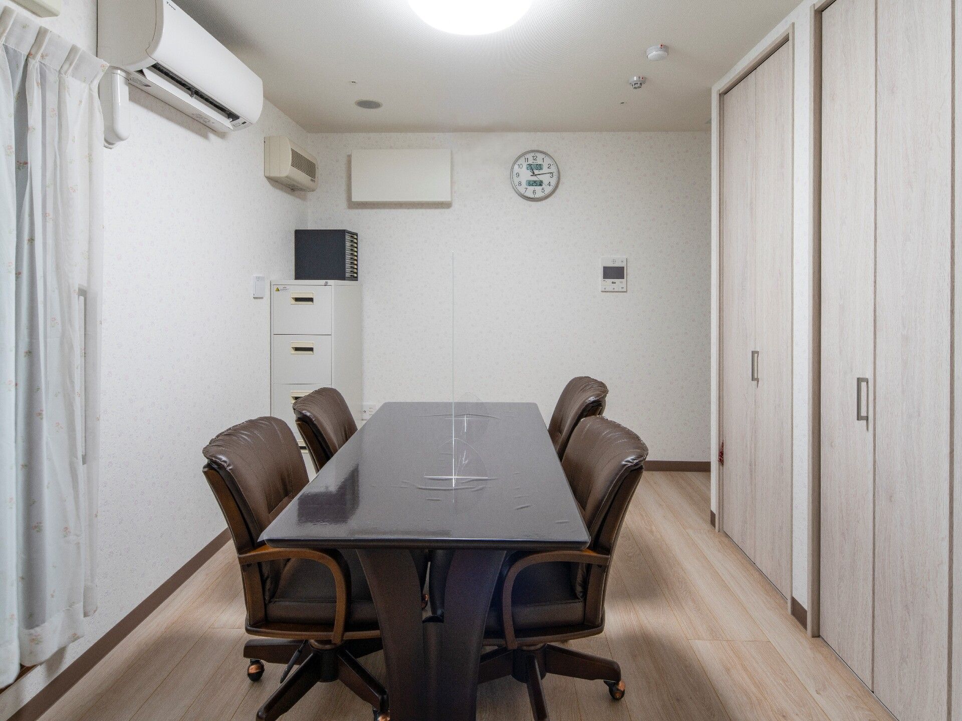 中央に肘置き付きの回転いすが4脚セットされたどっしりとしたテーブルが置かれ、壁にはエアコンや時計が付き、窓にはカーテンが付いている。