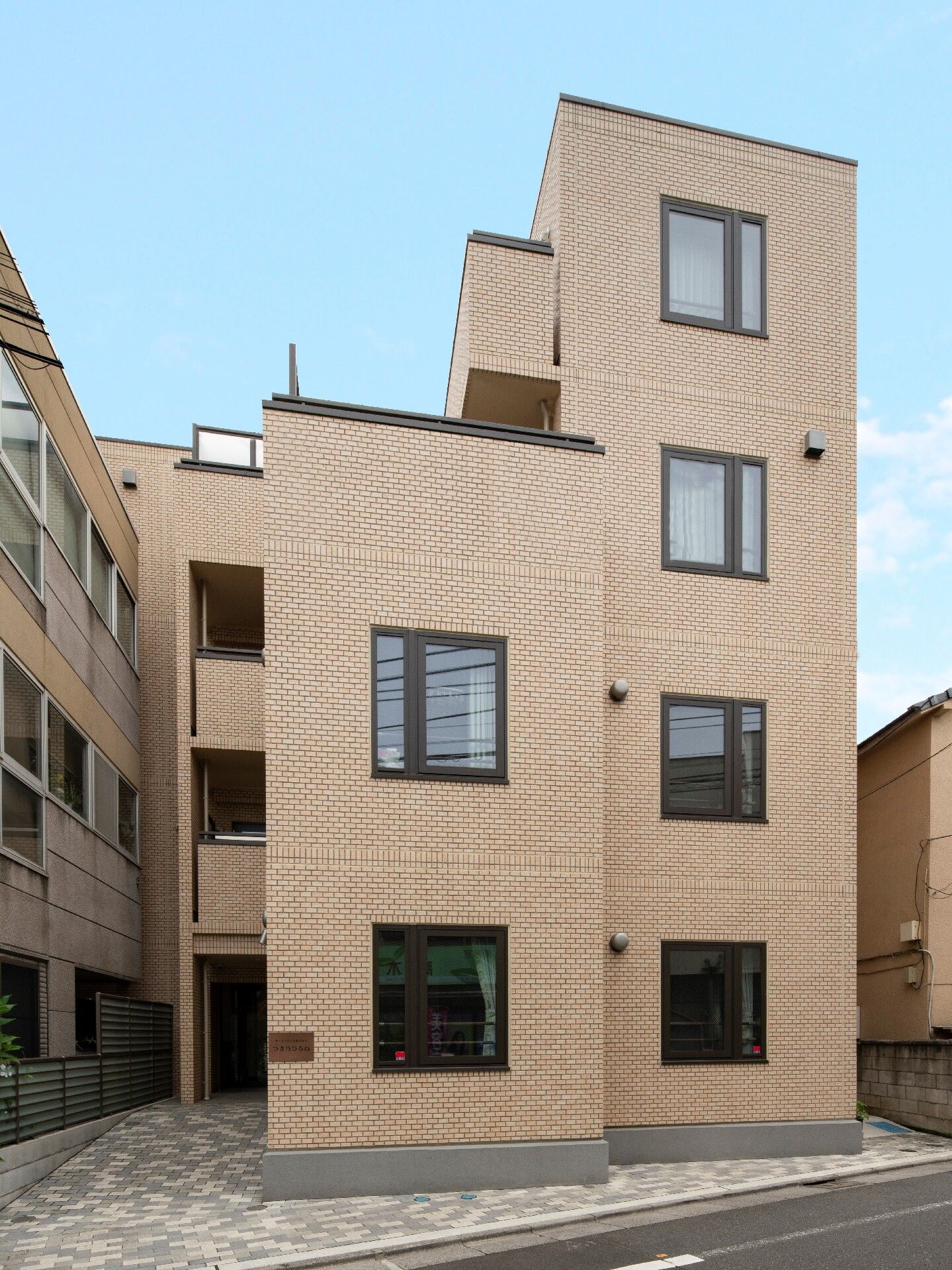 ベージュ系の建物は一部4階建ての3階建てで、たくさんの窓があり、道路に面している。空は青く、となりにも建物が建っている。