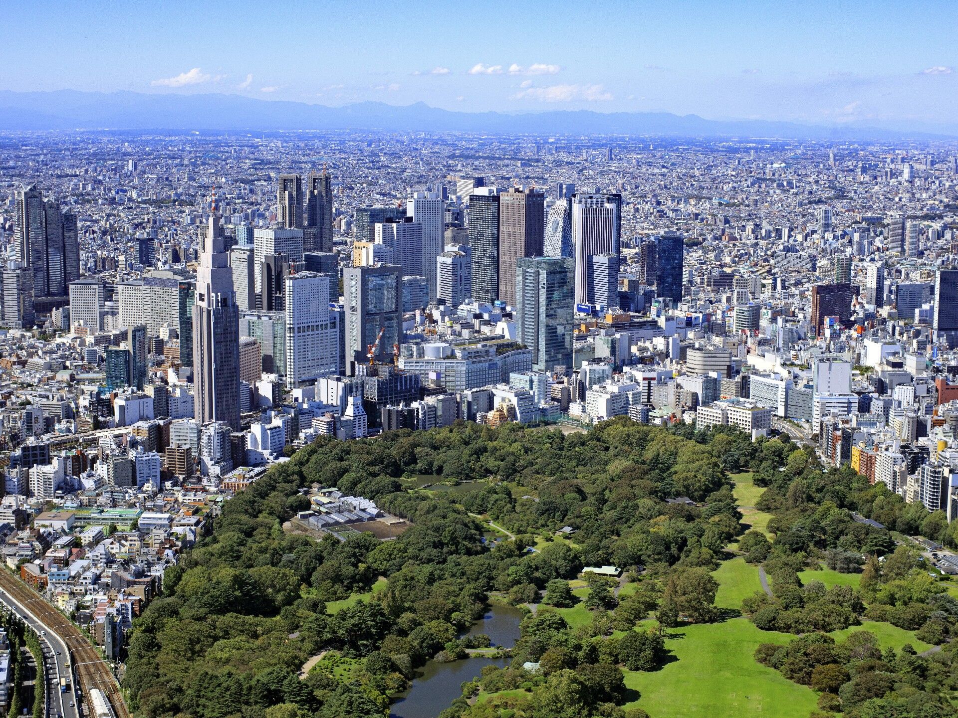 背の高いビルが建ち並ぶ街中の風景。手前には緑あふれる公園があり、奥にはうっすらと山が見える。