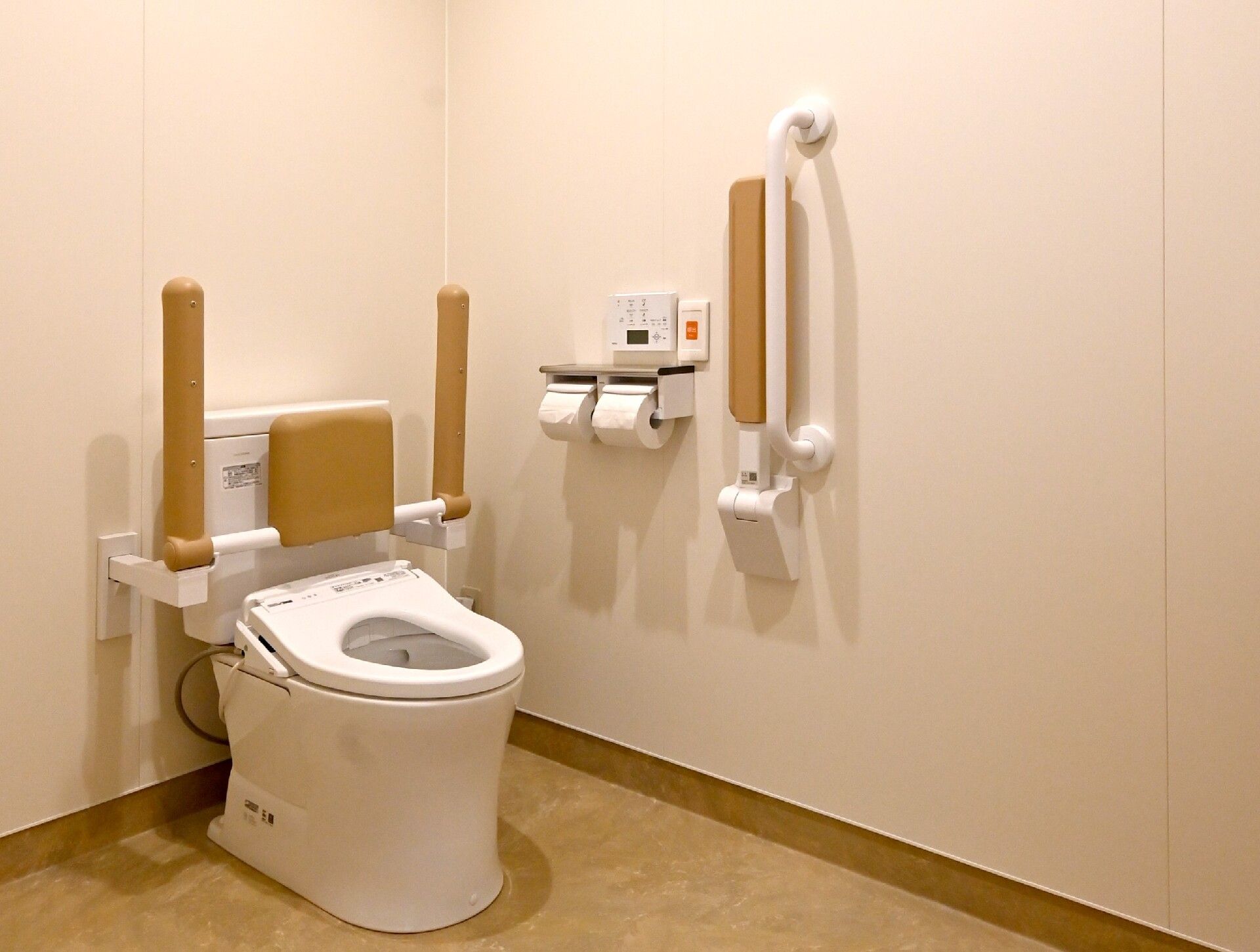 トイレの両サイドと壁にはベージュの手すりが付き、便座には背もたれもある。呼び出しボタンが付き、安全に配慮された造り。