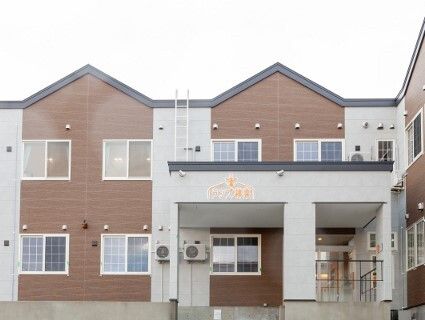 ブラウン系の2階建ての建物。玄関には住宅名がオレンジの文字で書かれた屋根が付いていて、窓も多い。