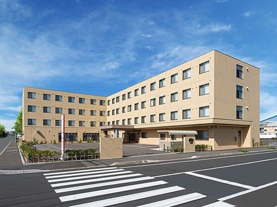 道路に面して建っているエル字型の建物はベージュ系の外壁で敷地内には駐車場があり、日当たり良好。