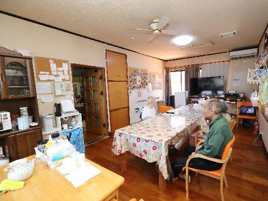 大きなテレビが置かれた食堂には花柄のテーブルクロスがセットされたダイニングテーブルが置かれ、入居者がいる。