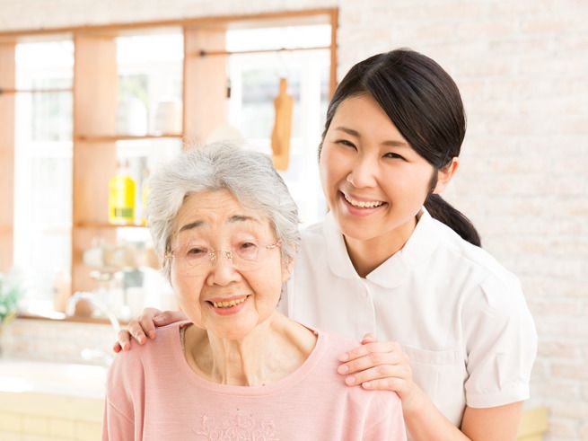 白衣を着た女性スタッフが笑顔で眼鏡をかけた高齢女性のそばにいて、両手を肩に置いている。