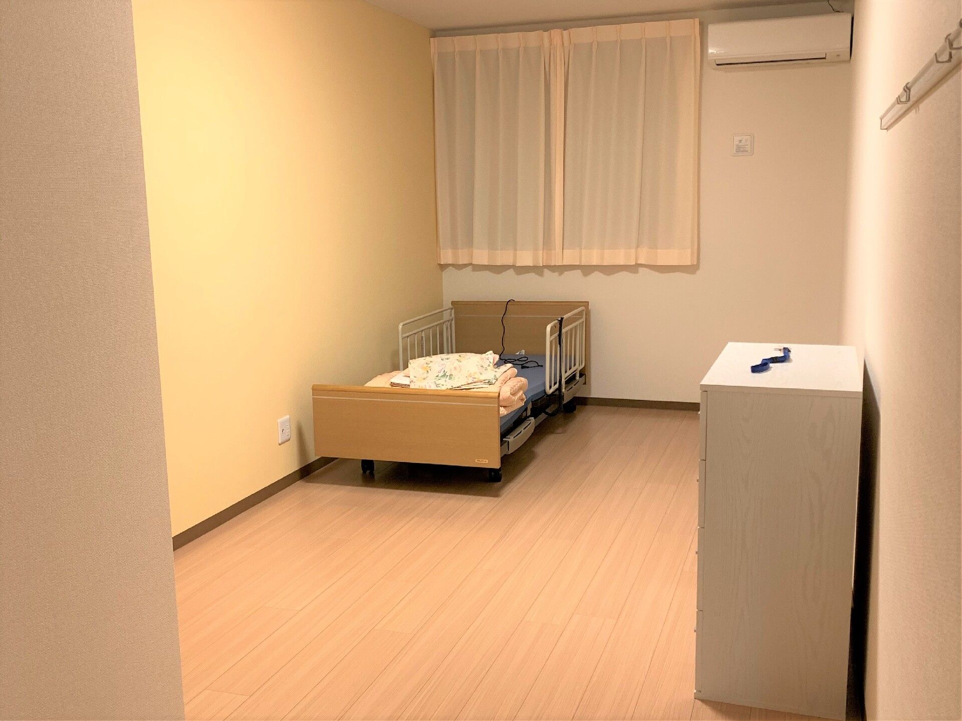 カーテン付きの大きな窓が付いたシンプルな居室にはベッド、タンスなどが付き、壁にはエアコンもある。