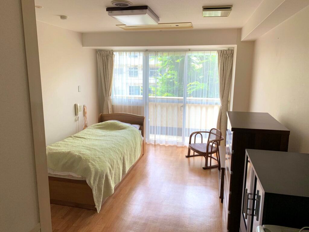 寝具付きのベッドやタンスなどの家具が置かれたフローリングの部屋には緊急通報装置があり、バルコニーも付いている。