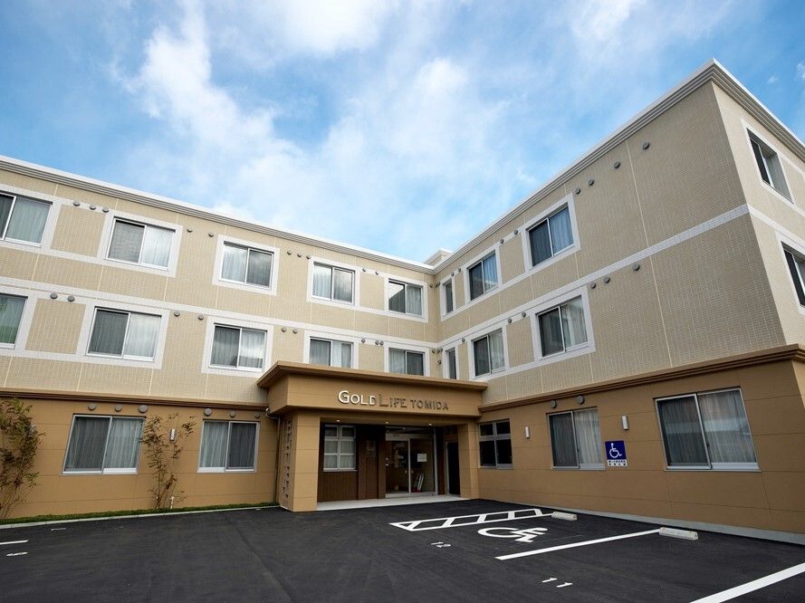 3階建ての建物はエル字型で1階部分が濃いベージュ、2階より上が薄いベージュ。駐車スペースもあり、玄関には屋根も設置されている。
