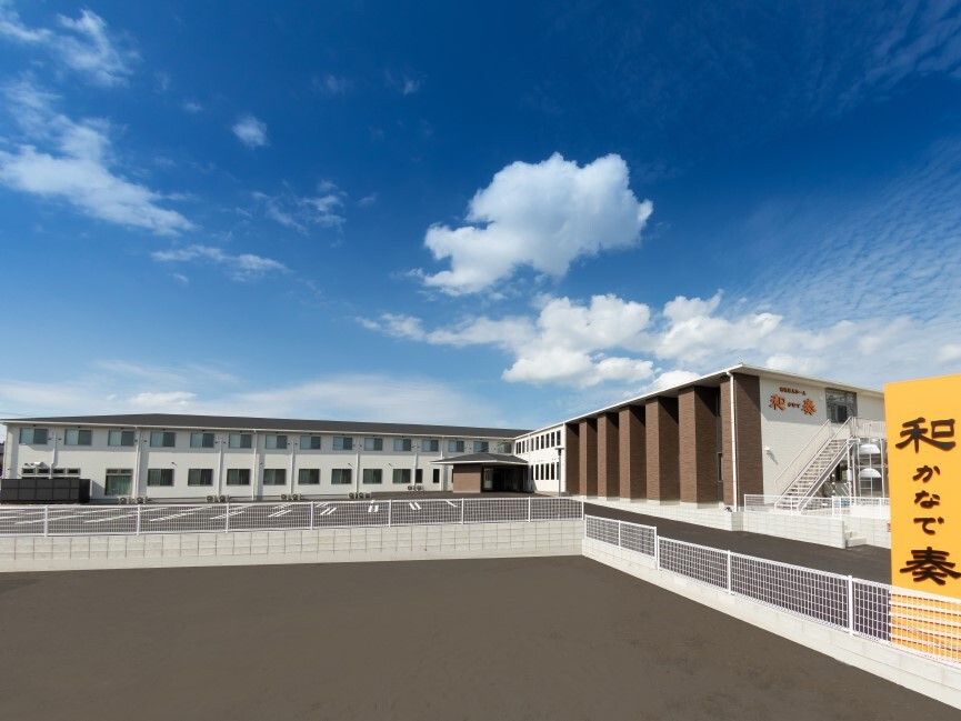 青空の下に建っている２階建ての建物はエル字型でホーム名が外壁に書かれ、大きな駐車場が付いている。