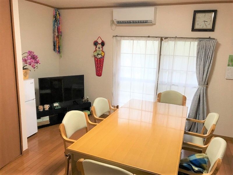 明るいベージュのダイニングテーブルには6脚の椅子がセットされ、周りにはテレビや冷蔵庫が置かれている。