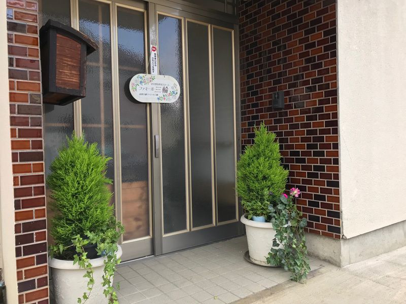 木製のポストやウェルカムボードが飾られて玄関。木や花が植えられたプランターも置かれている。