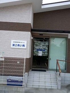 ブラウン系の建物の玄関先には手すり付きの階段が付いている。外壁にはホーム名が書かれたプレートもある。