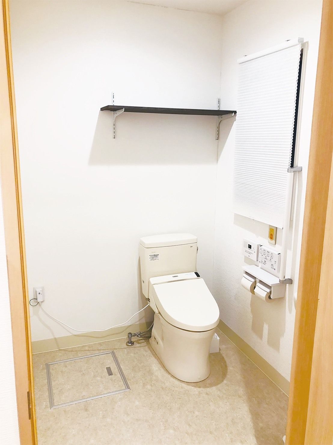 オレンジの呼び出しボタンが付いた広いトイレ。壁には操作ボタンやトイレットペーパーホルダーもあり、便座の上部には棚も設置されている。