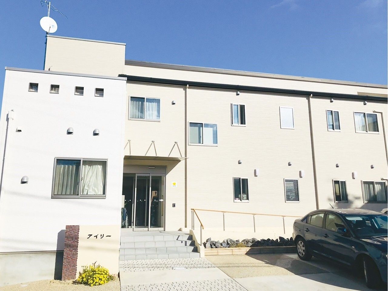 ホーム名が書かれた館銘板が設置され、２階建ての建物の玄関には手すりが付いたスロープと階段があり、駐車スペースには車が止まっている。