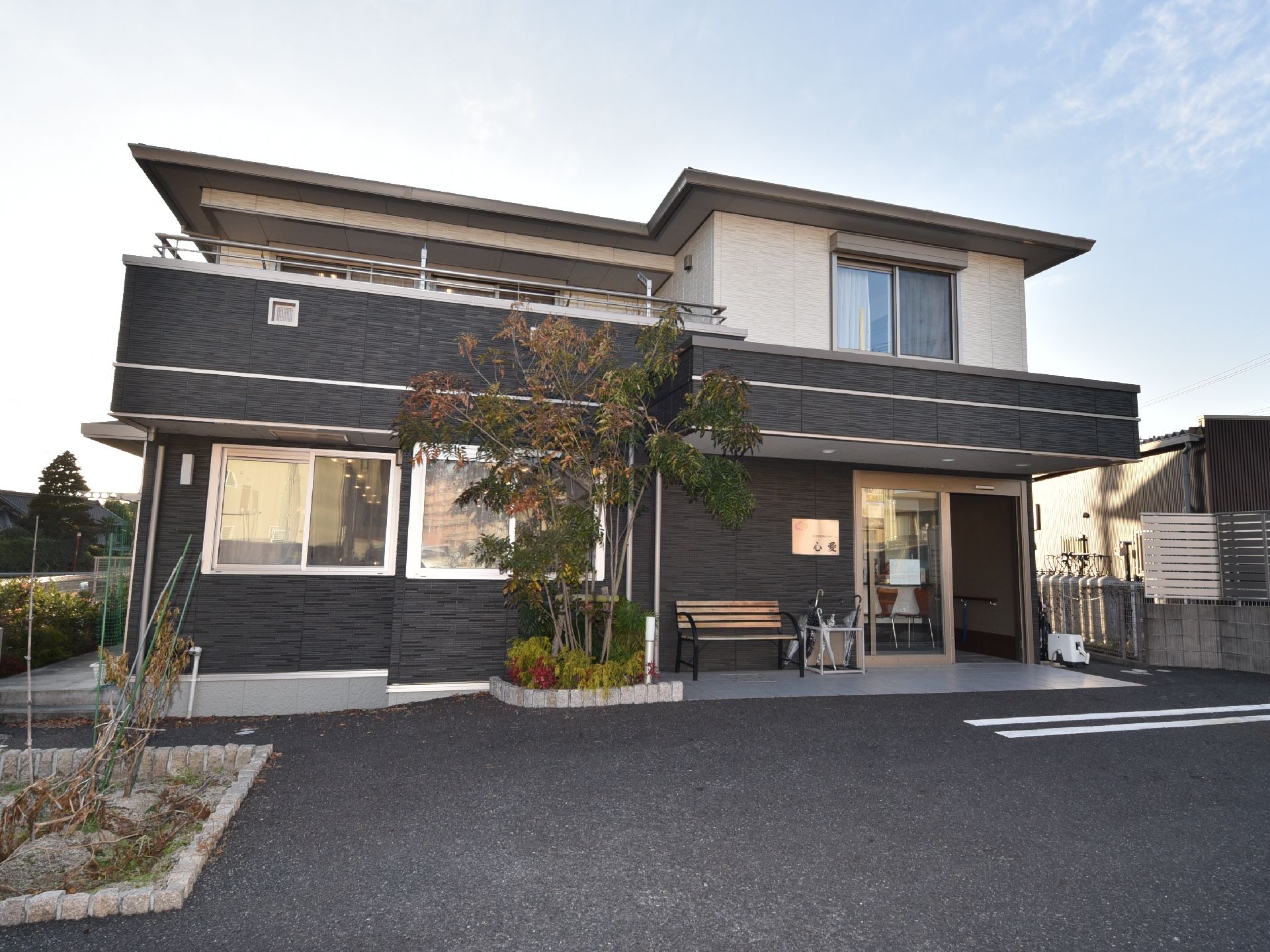 ダークグレーと白の２階建ての建物は玄関に屋根があり、木製のベンチが置かれ、駐車場もある。