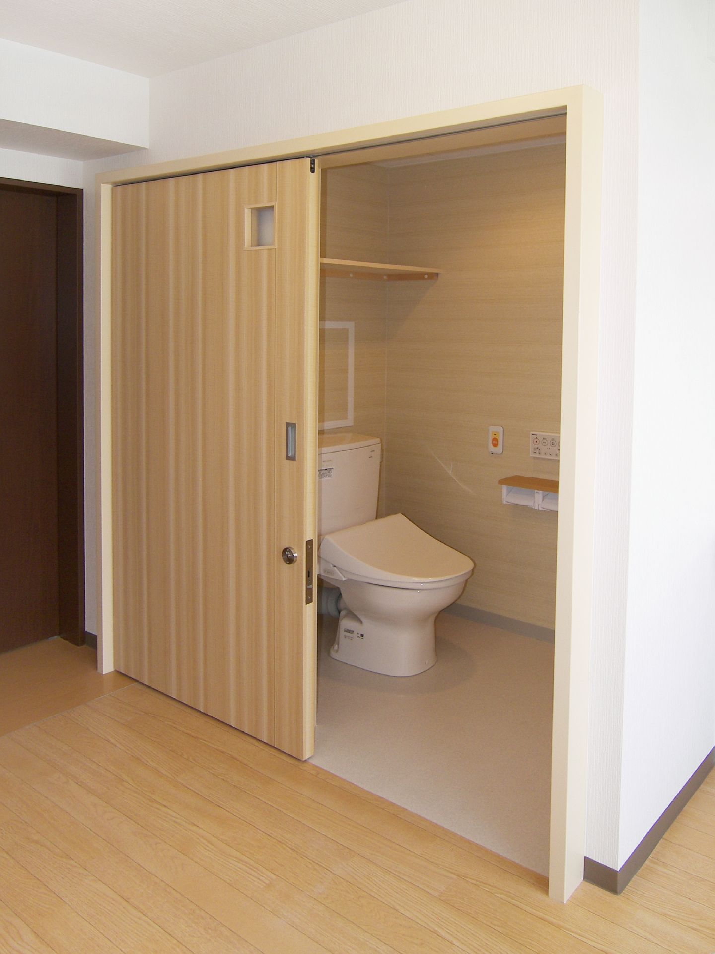 大きなベージュのスライドドアが付いたトイレ内は広く、オレンジの呼び出しボタンやトイレットペーパーホルダー、操作パネルなどが壁についている。