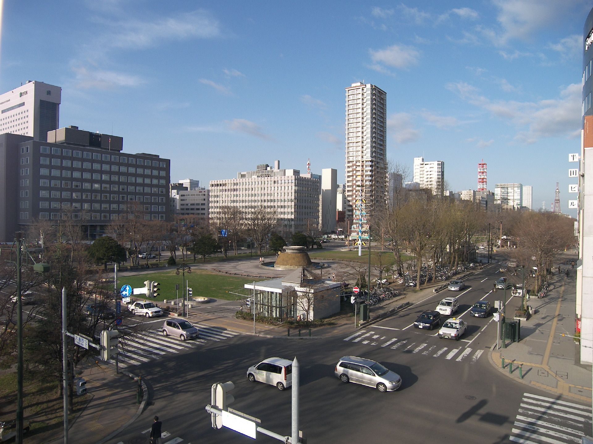 十字路の交差点が目の前にあり、車が数台行き交っている。奥には背の高いビルが建ち並んでいる区域があり、空には青空が広がっている。