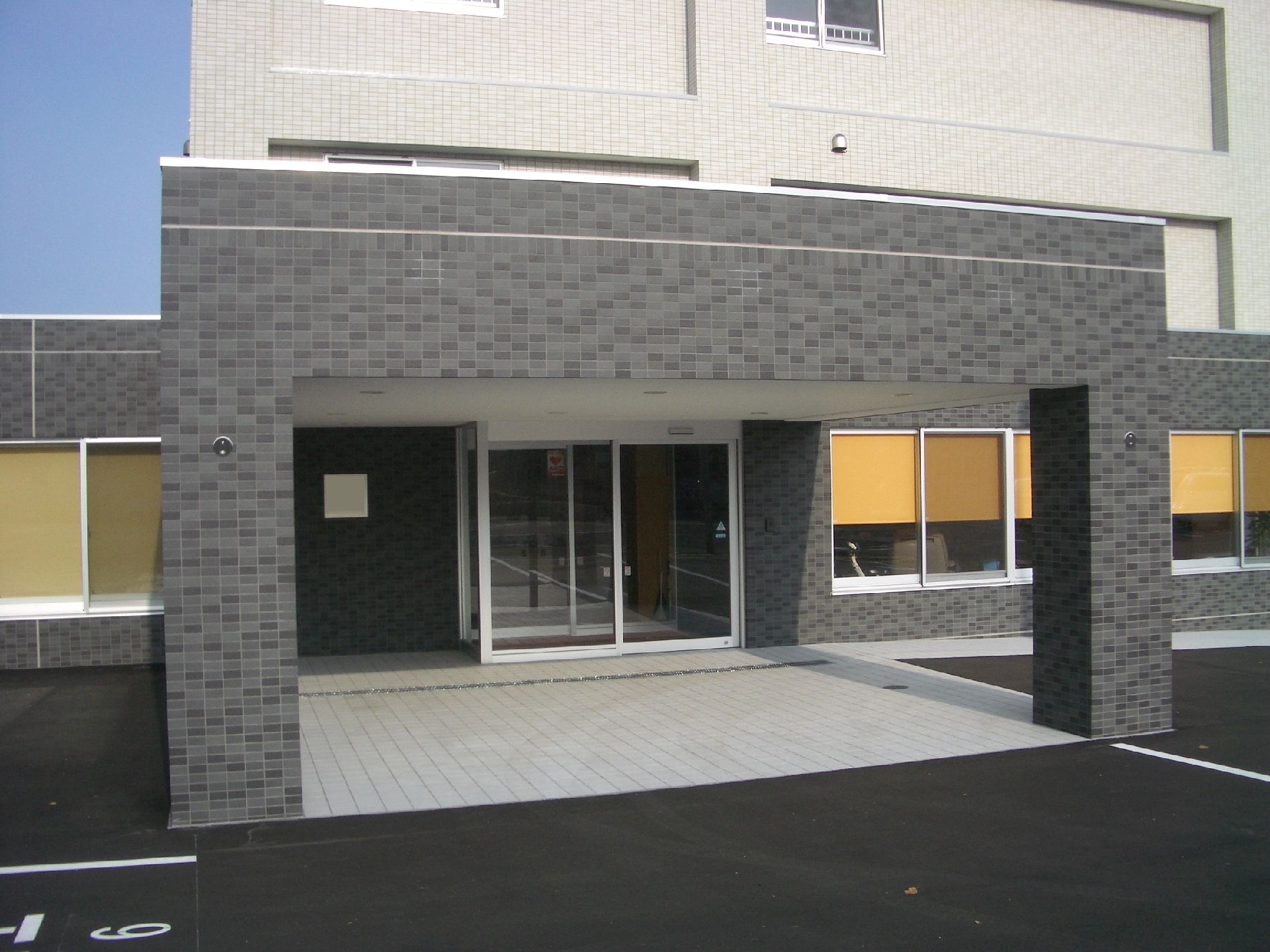 白とグレーの重厚感のある建物。玄関先には大きな車寄せが付き、段差がなく、出入り口は自動ドア。敷地内には駐車場もある。