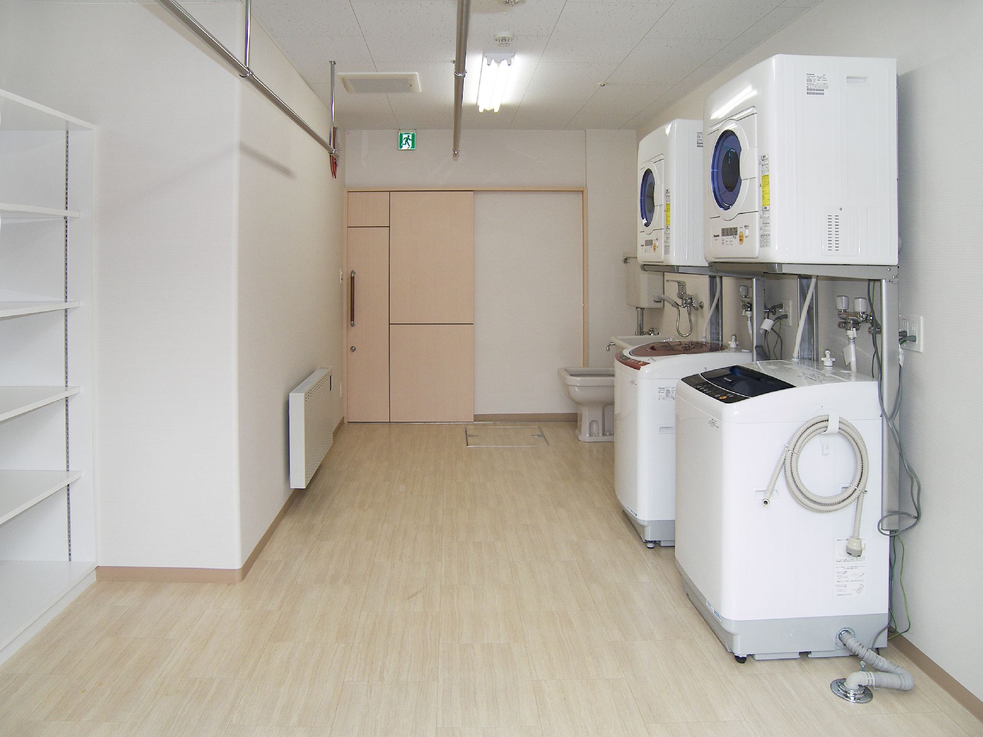 白い縦型の洗濯機と乾燥機がペアで置かれている。白い棚には何も置かれておらず、壁にはパネルヒーターも付いている。