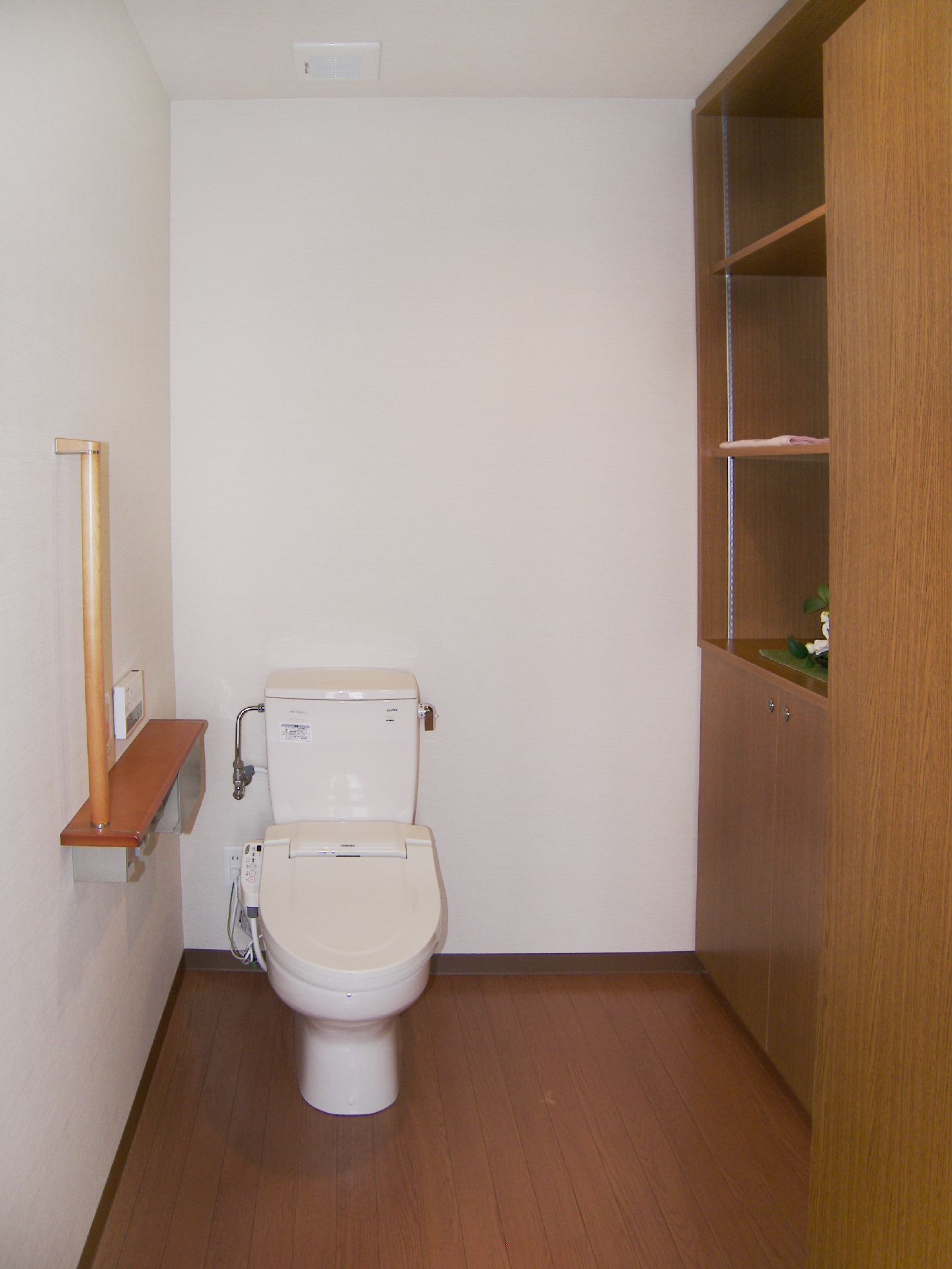 広いトイレには手すりや呼び出しボタンがあり、右側には収納棚が設置され、白い花が飾られている。